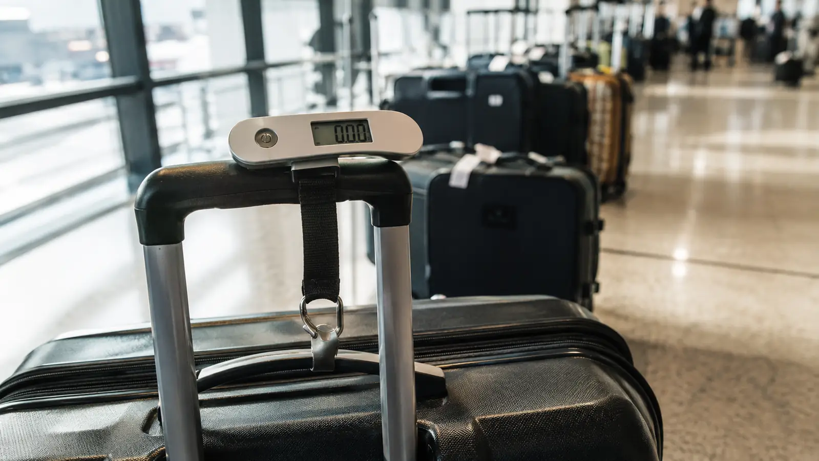 A small handheld luggage scale resting on the handle of a suitcase beside a tidy row of checked bags in a bright terminal, natural light