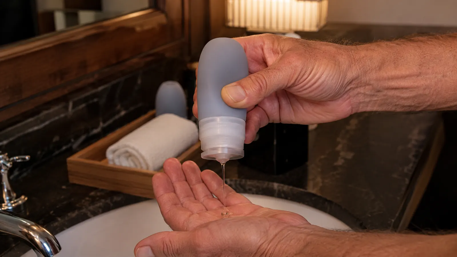 A hand squeezing gel from a small soft silicone travel bottle over an open palm beside a hotel bathroom sink