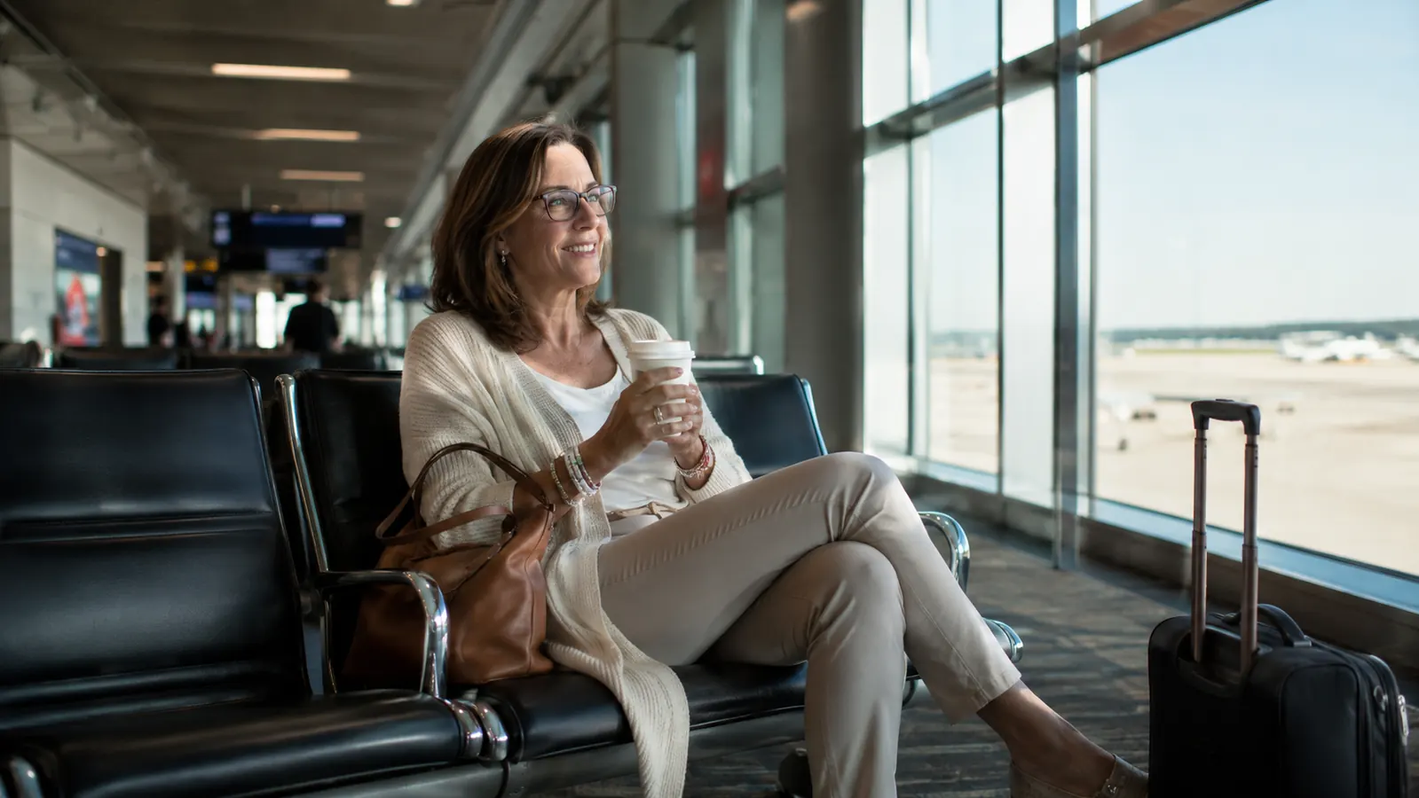 A woman in her fifties sits at a tall airport window in soft morning light, a small suitcase and a tote on the floor beside her, looking out toward the tarmac
